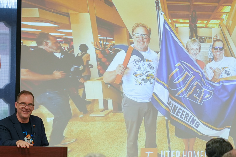 Individual stands at a podium during a UTEP Engineering homecoming event while presenting a slide featuring a homecoming photograph and UTEP Engineering flag, with attendees seated in the foreground and a large projection screen visible behind.